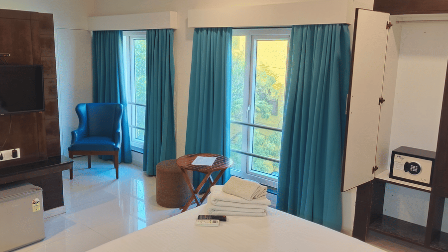 A room interior showing large windows with teal curtains, a flat-screen TV, a mini-fridge, and an open wardrobe with an electronic safe at The Bull Boutique Hotel.