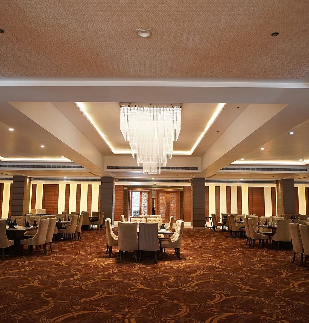 A large and sophisticated event space at Nirvana Luxury Hotel, Ludhiana, with round tables and chairs arranged on a patterned carpet under a grand chandelier and recessed lighting.