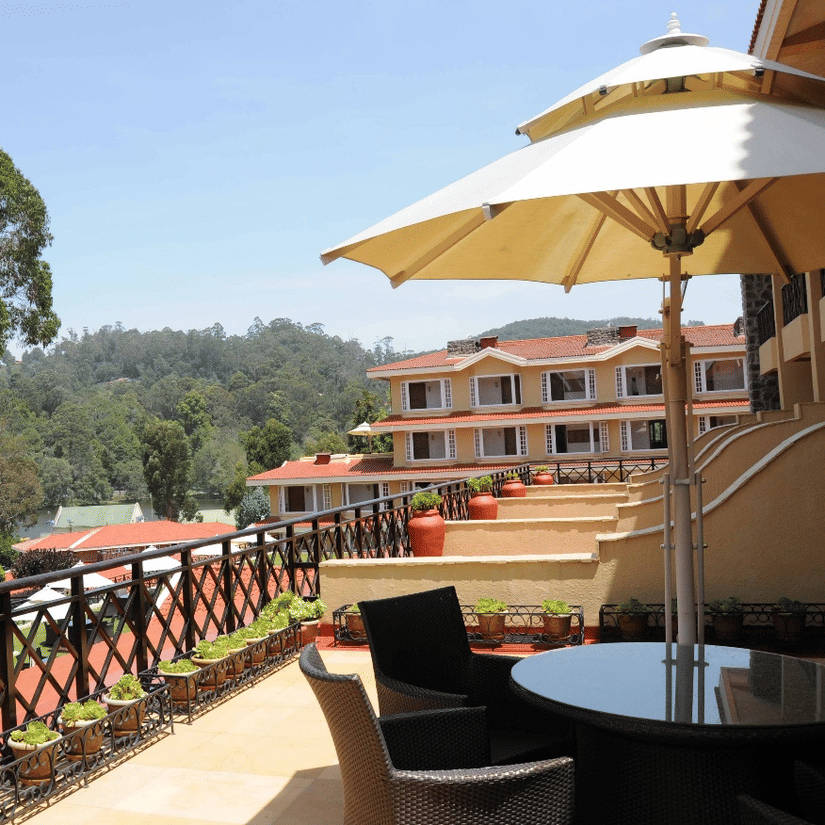 View of the terrace at The Carlton Kodaikanal with outdoor seating, sun umbrella, wooden railing, and a serene lake and hillside beyond.