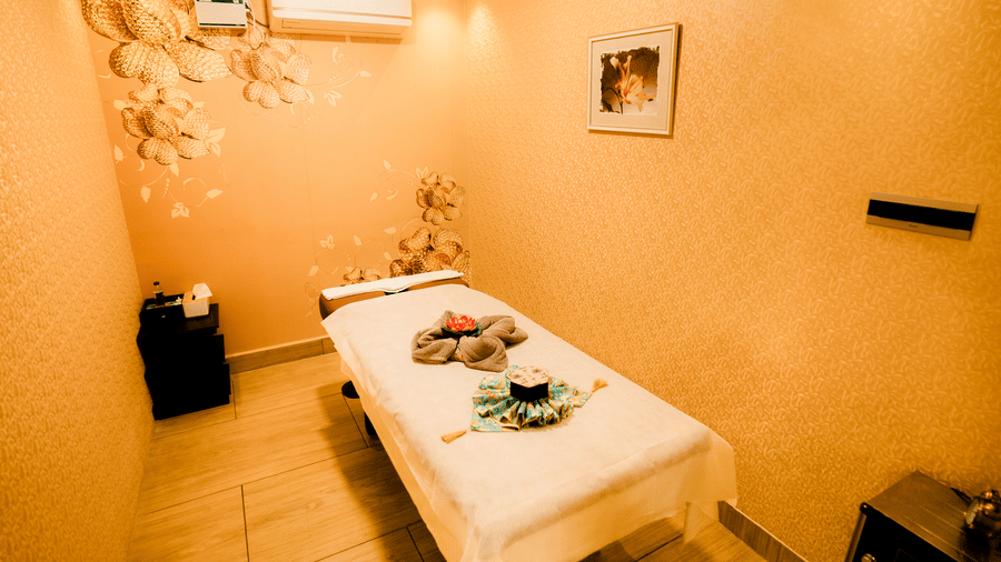 Spa treatment room with a massage bed, towels folded into a flower, gold floral wall decor and framed art at Hasta Spa, Ilara Hotels & Spa, Tada.