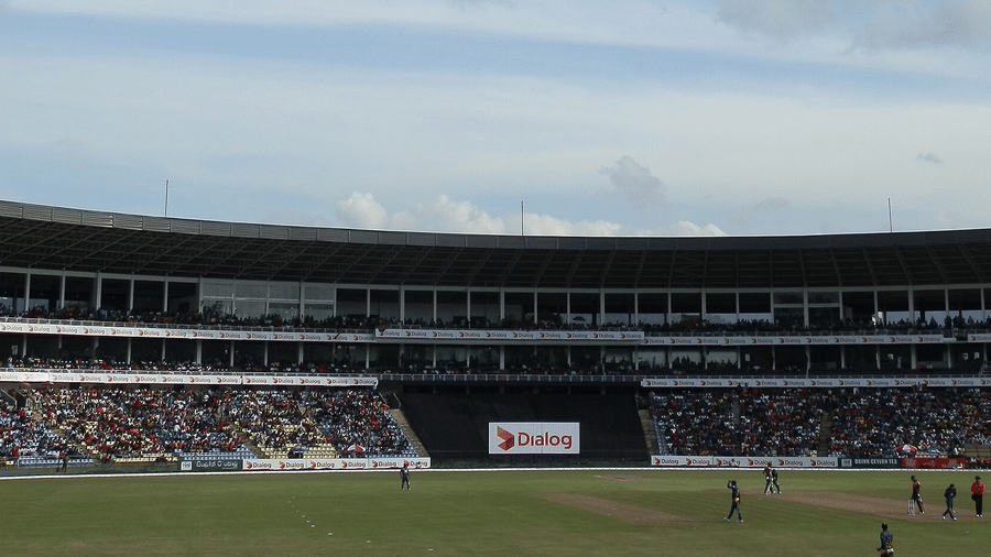 Wide-angle shot of cricket arena showing main pavilion, commentary boxes, and grassy playing area under soft daylight.