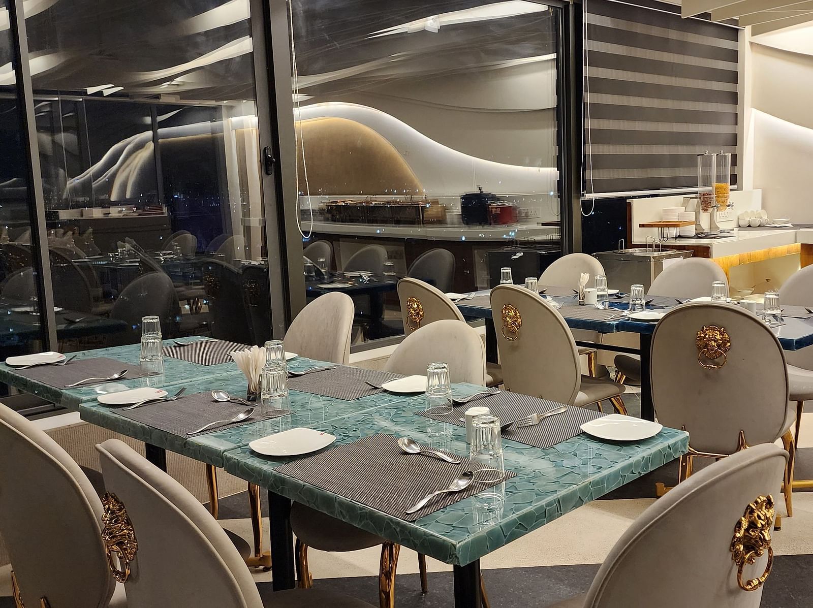 An elegant dining area with white and gold chairs and set tables, located beside large windows offering a view outside.