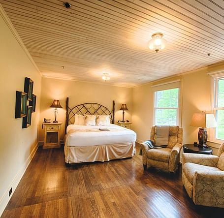 full view image of a room with wide windows featuring a cozy double bed and comfortable cushioned armchairs in front of it - Historic Tapoco Lodge