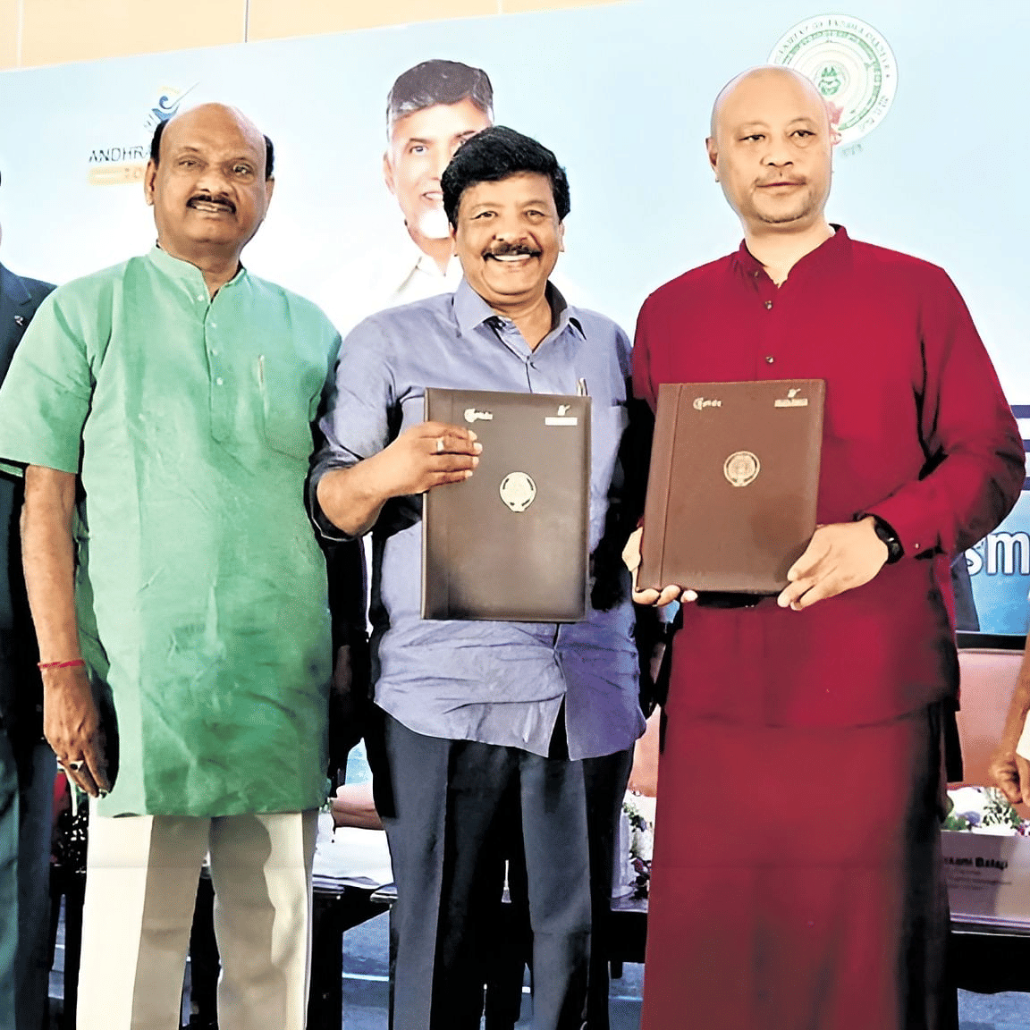 A group of dignitaries at a formal event holding signed documents, posing for a photo. A backdrop with official logos and portraits is visible behind them.