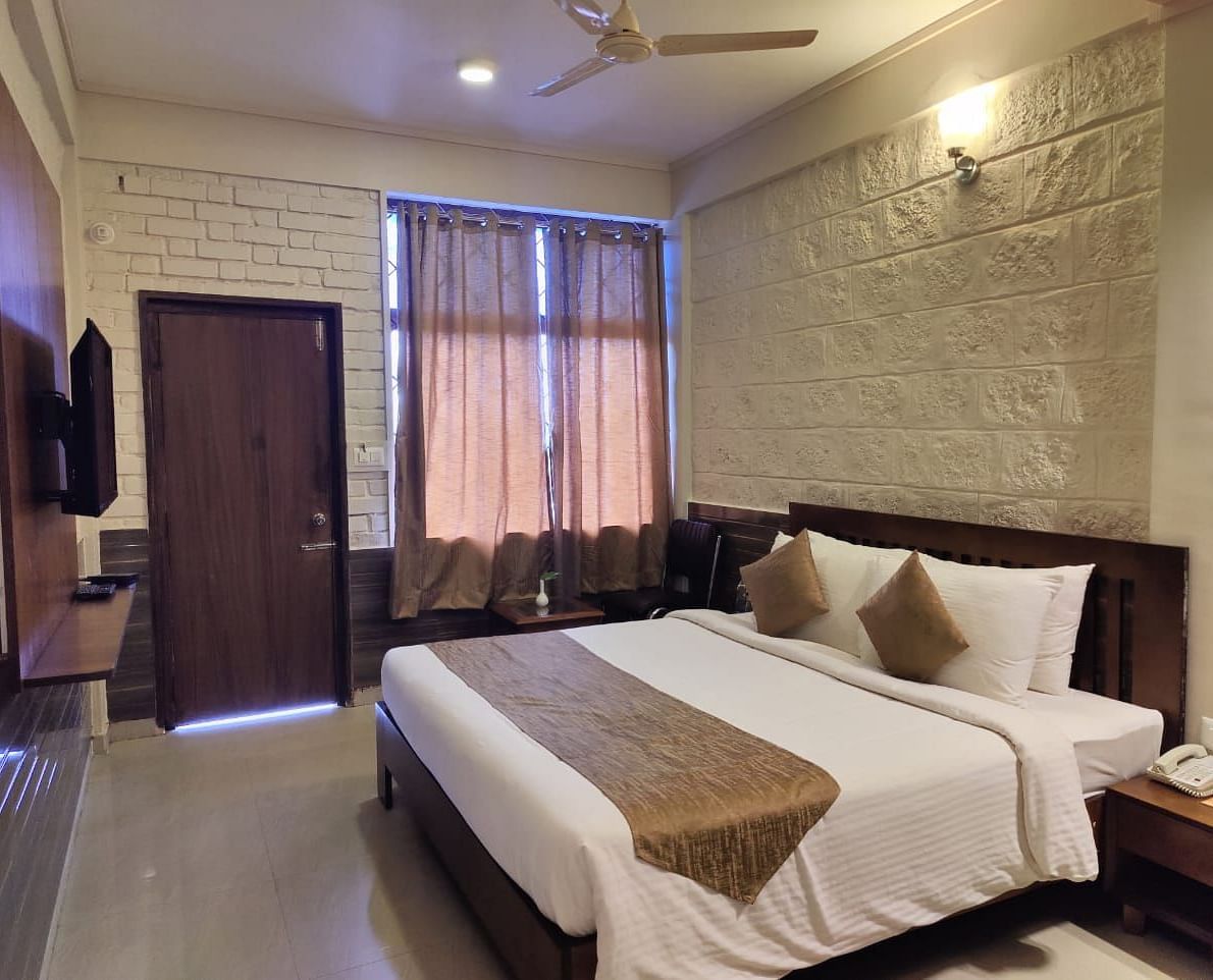 Bedroom with a huge bed, bedside table, windows with curtains, wooden door, and a TV | Standard Room | Woodstock Resorts, Coorg