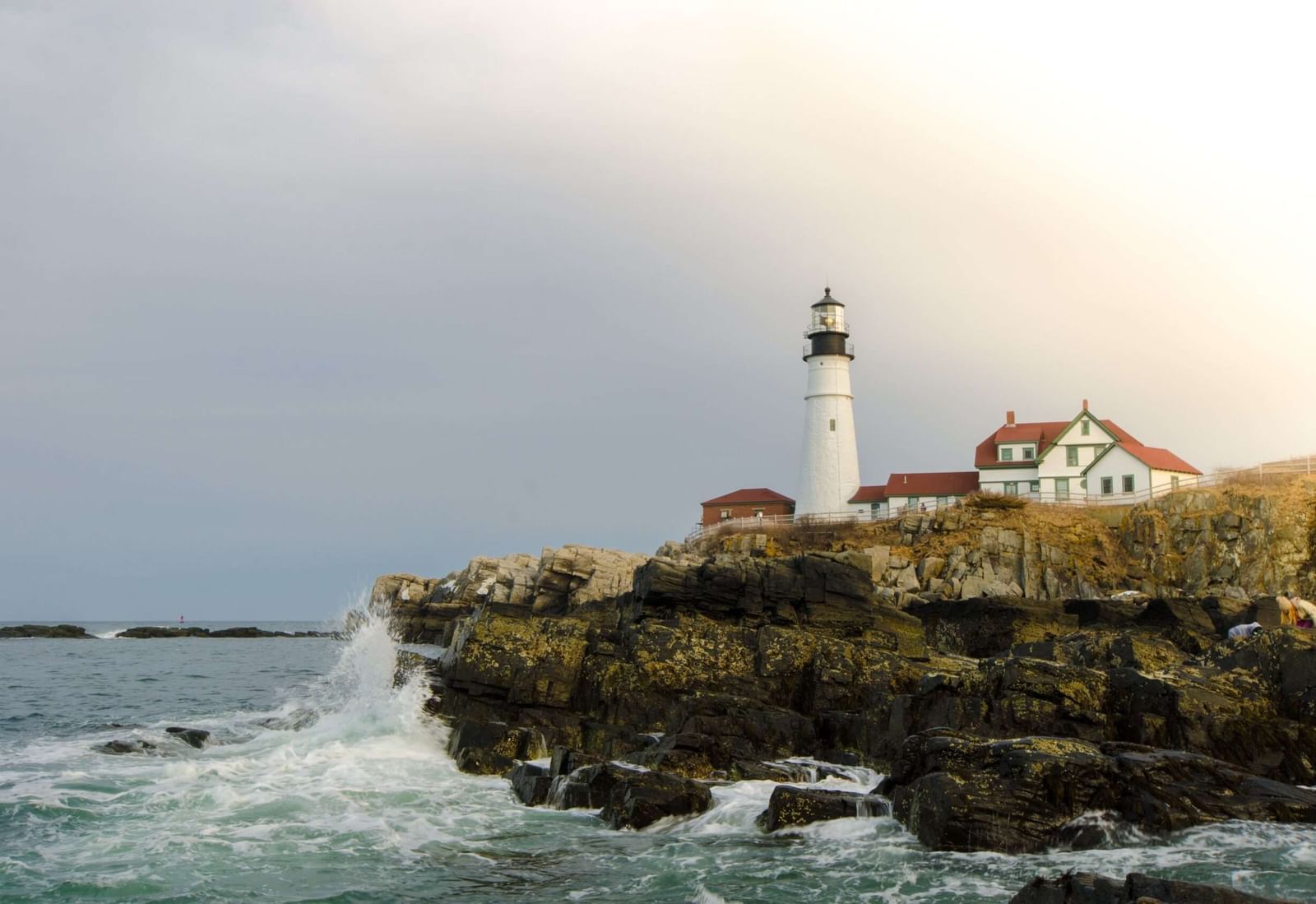 A wide angle shot of a light house at the end of a cliff with waves crashing against the shore under a stormy sky.