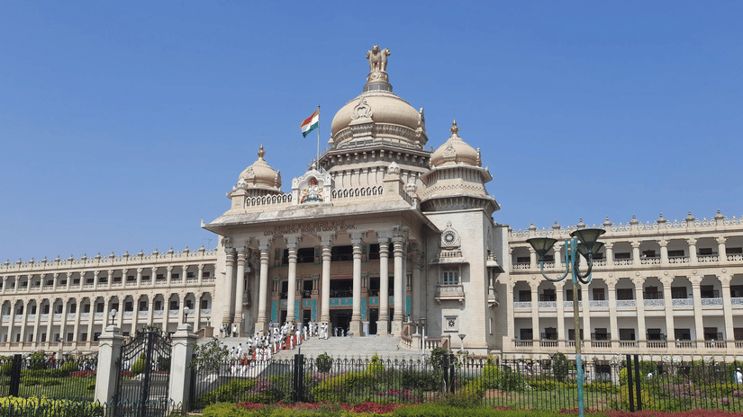 The impressive stone facade of Vidhana Soudha stands beneath a clear sky with grand pillars, detailed domes, and the national flag flying high.