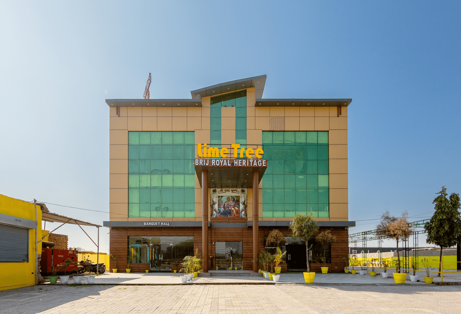 A daytime view of Lime Tree Hotels & Banquet, Vrindavan building with glass panels, yellow accents, and an open parking area in front.