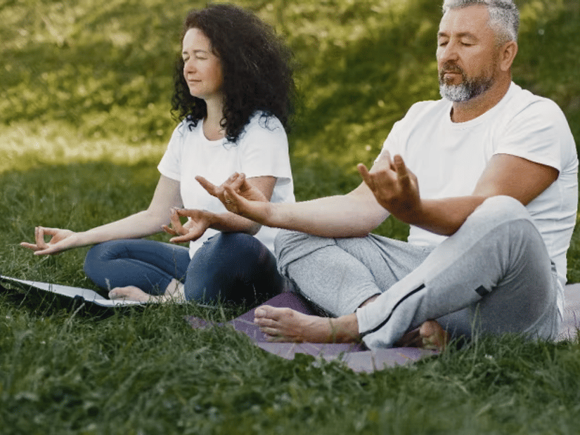 A man and a woman seated cross-legged on a mat on the grass, meditating with eyes closed