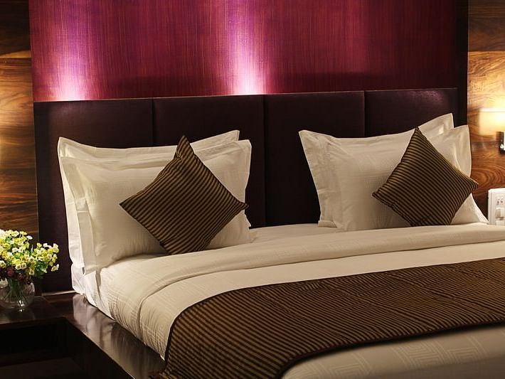 A warmly lit hotel room showcases a king size bed with a prominent dark headboard, crisp white linens, and decorative throw pillows.
