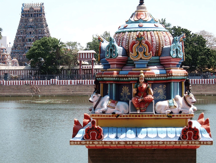 A colorful temple chariot or float on a body of water with a temple tower in the background.
