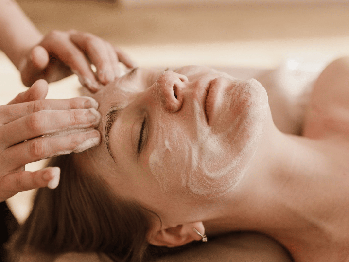 A person with their eyes closed is receiving a facial treatment, with a creamy substance being applied to their face