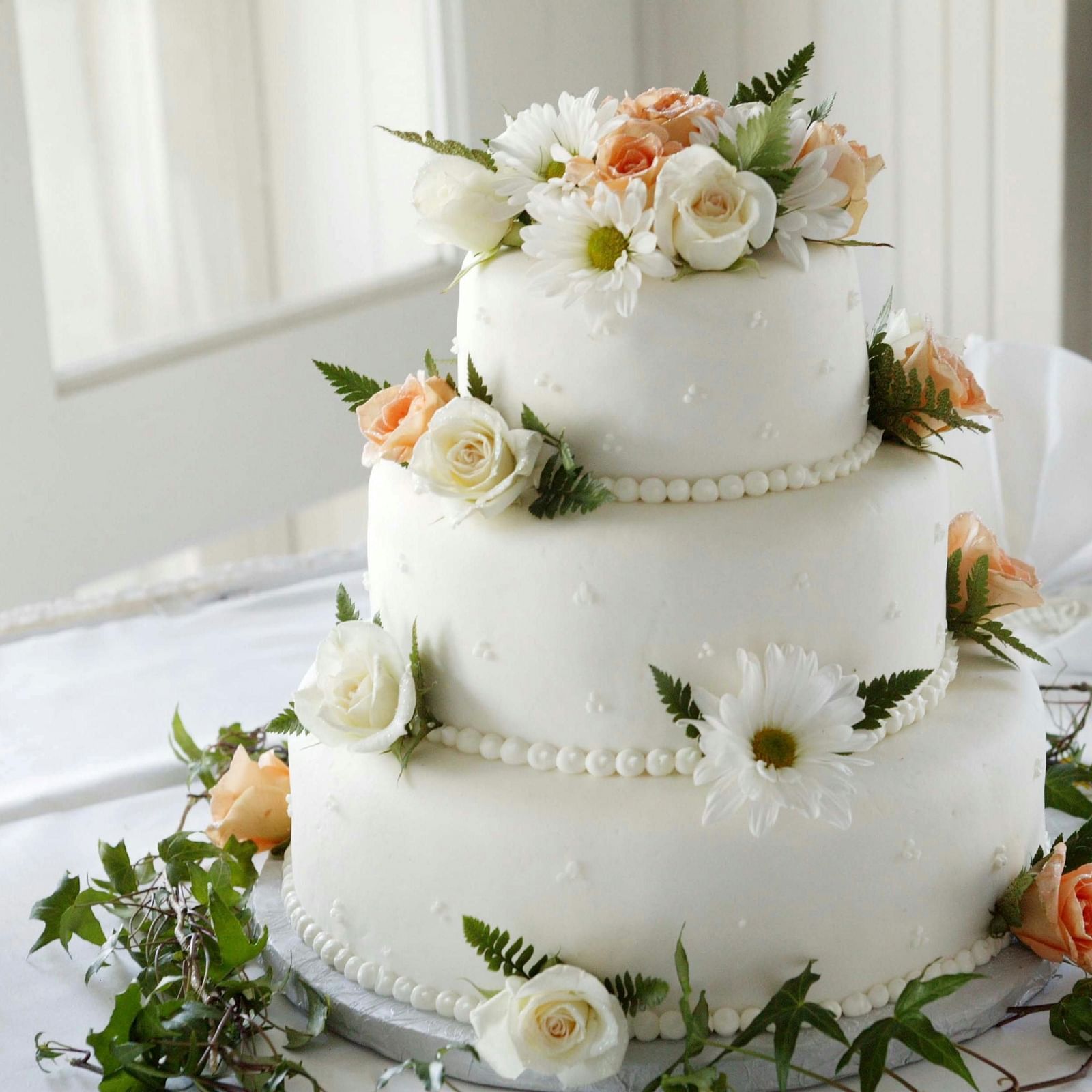 A three-tiered white wedding cake adorned with fresh flowers and greenery, set on a table with a white cloth