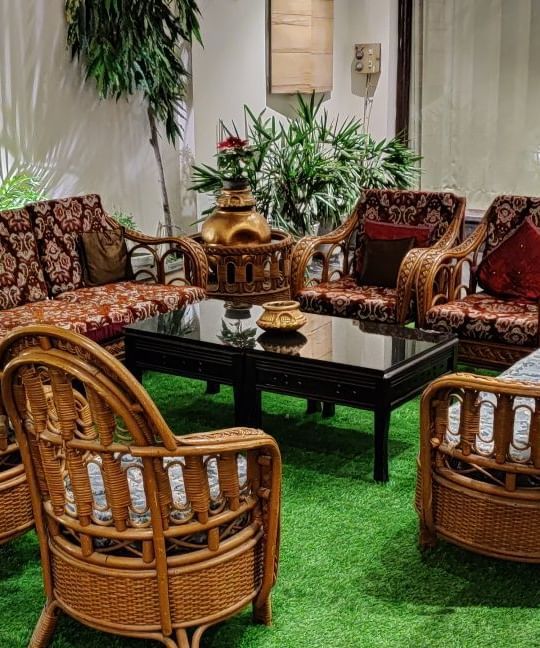 An interior shot of a lounge or seating area at Hotel Exotic Amritsar, featuring a set of wicker furniture including a sofa, chairs, and a table, all arranged on a green rug. 
