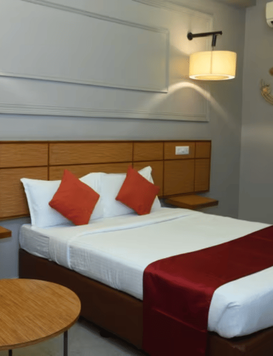 Bedroom with double bed, red runner, pillows, green chair, table and wall lamps at Max Hotel Prayagraj.