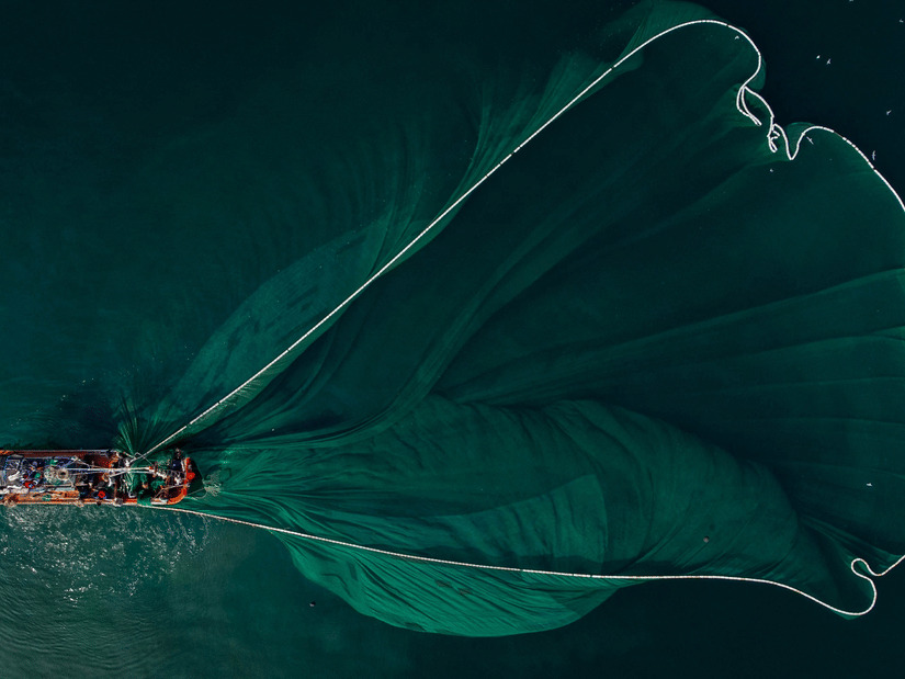 An aerial view of a small fishing boat on dark teal water, trailing a translucent fishing net that fans out across the surface.