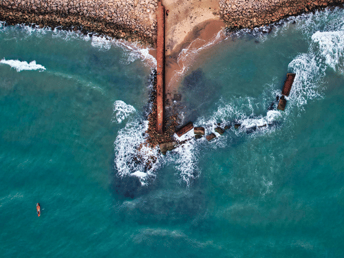 High-angle shot of the ocean waves crashing against a dark rocky pier and sandy shoreline.