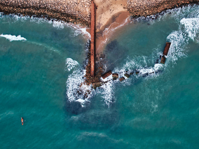 High-angle shot of the ocean waves crashing against a dark rocky pier and sandy shoreline.