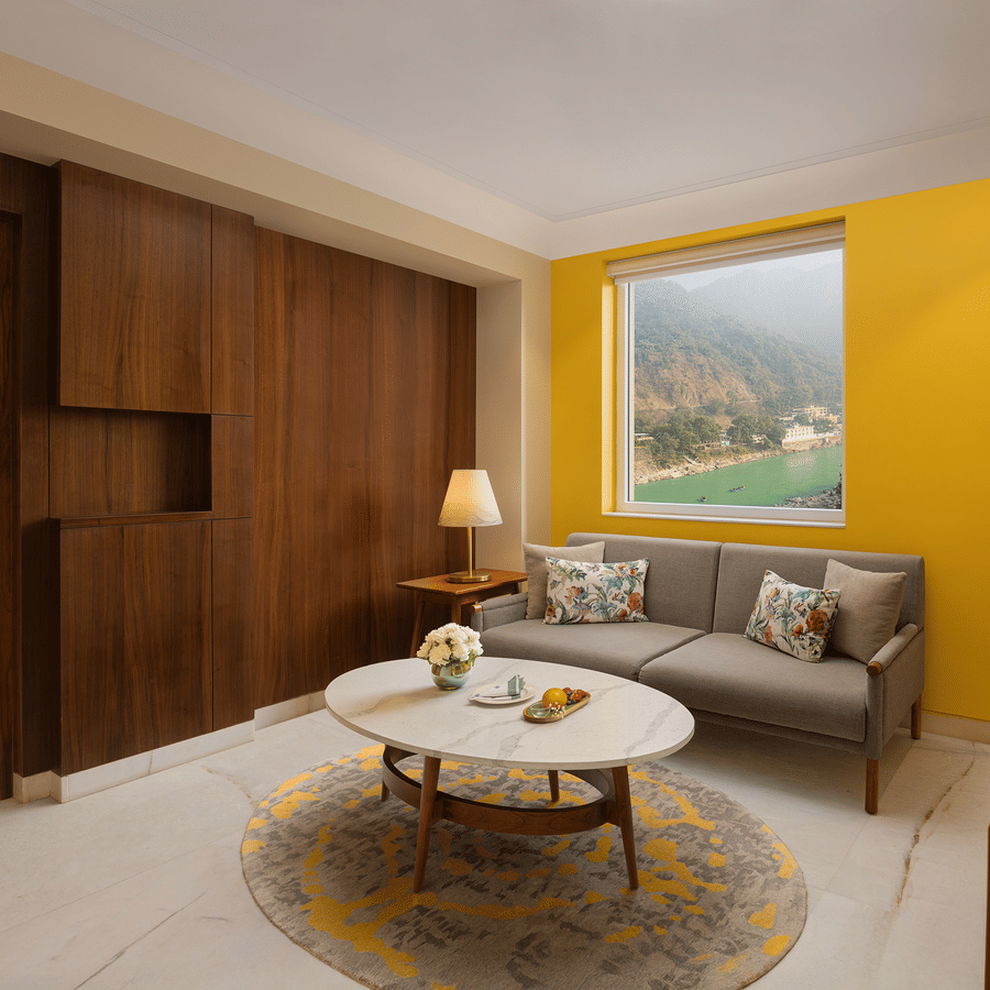 Modern hotel living area with a grey sofa, yellow accent wall, and a large window overlooking a river and mountains.