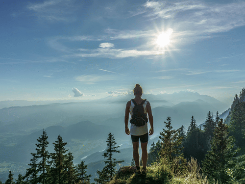 A person standing on a hilltop overlooking layered mountain ranges and trees under a bright sky.