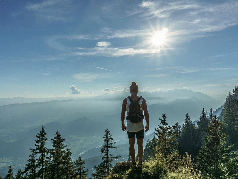 A person standing on a hilltop overlooking layered mountain ranges and trees under a bright sky.
