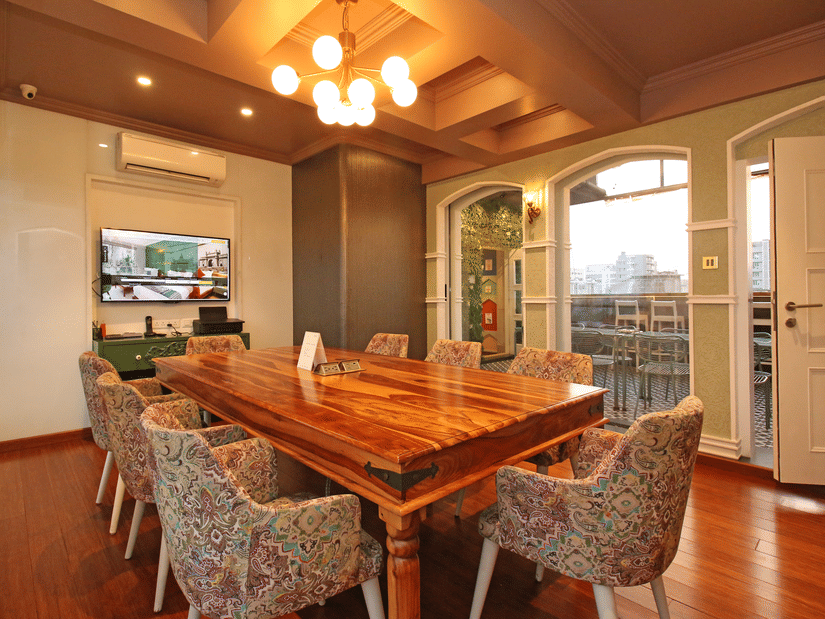Warm, stylish dining and meeting space at Theory9 Premium Service Apartments, Bandra, featuring a long wooden table, patterned chairs, soft lighting, and large windows with city views.