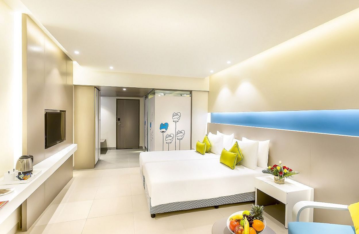 A spacious room features a large bed with a headboard stretching across the wall. Cushion chairs sit beside a wooden bedside table, and a wall-mounted television is positioned opposite the bed