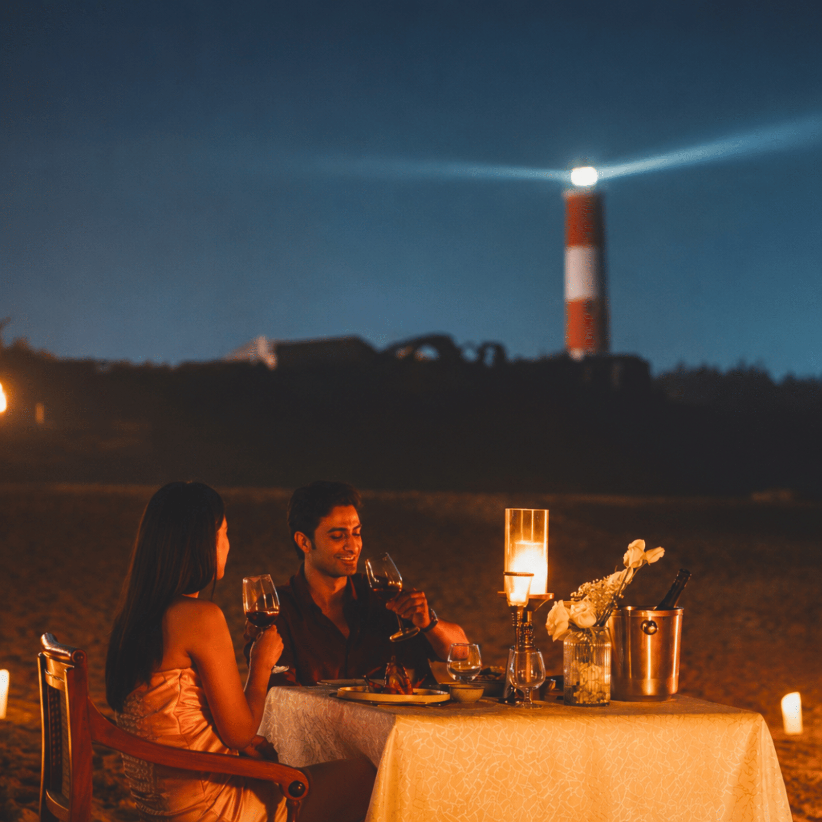 Two people at a candlelit dinner table on the beach at night, with a lighthouse beam behind, at MAYFAIR Palm Beach Resort, Gopalpur-on-Sea.