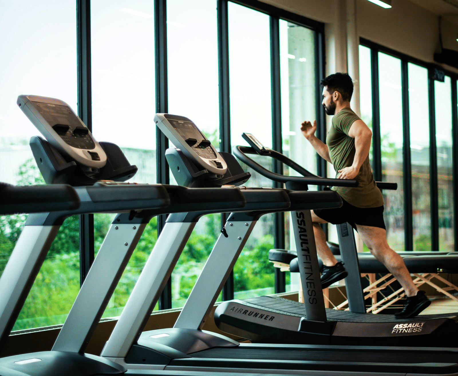 A man with a beard is running on a treadmill in a modern gymnasium with large floor-to-ceiling windows overlooking green trees.