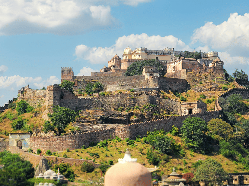 Panoramic view of Kumbhalgarh Fort spread across a green hillside, with ancient stone walls, rugged terrain, and white clouds overhead