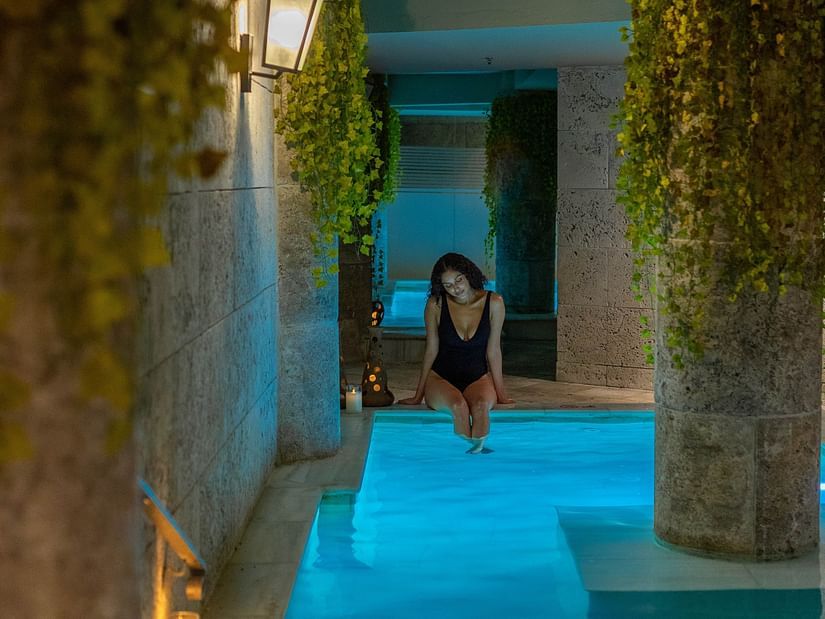 Woman relaxing by an indoor pool in a serene spa setting, surrounded by stone walls, soft lighting, and hanging green plants. 