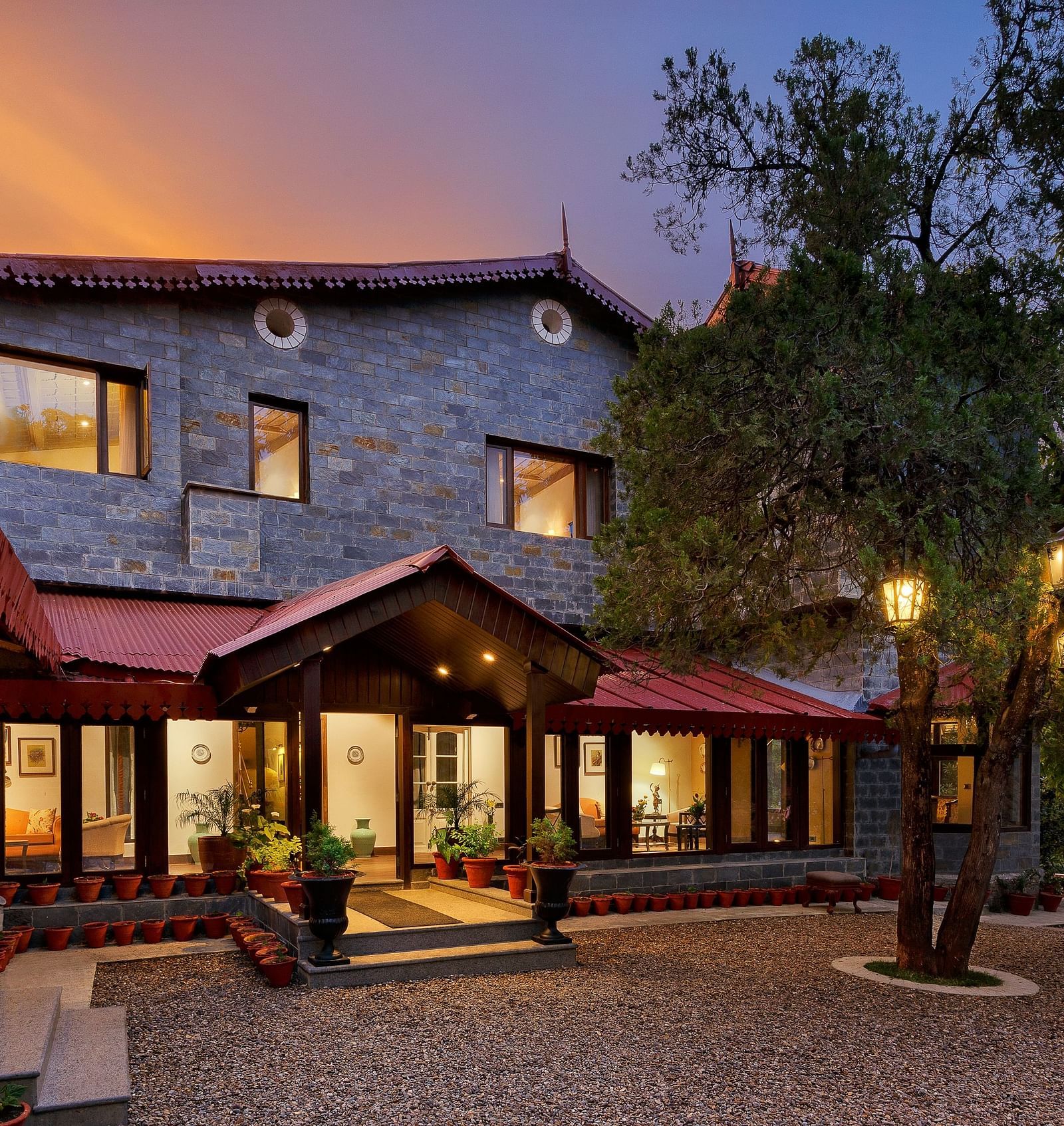 An Image of facade of  building with a lot of wooden details, which stands behind a Twin tree with lights, captured in the photo after the golden hour - The Naini Retreat