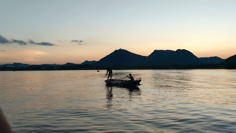 2 people sailing on a small boat on a lake during sunset.