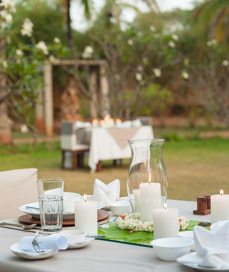 An outdoor dining setup with white tablecloths and chairs, green lawn and trees in the background - Shreyas Retreat