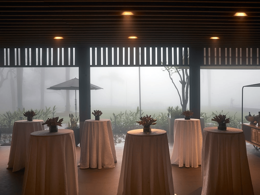 Indoor event space with high cocktail tables draped in white linen, each holding a floral centerpiece, looking out through large windows onto a foggy, misty garden.