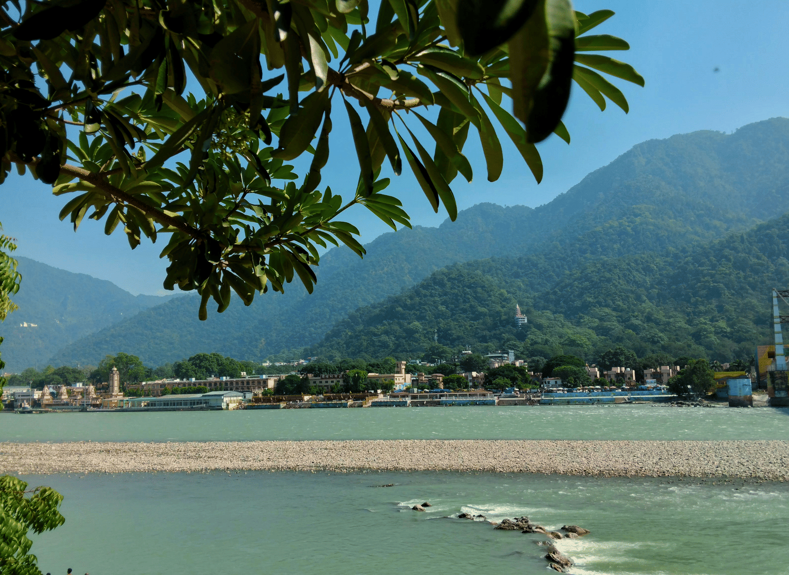 A scenic view of a river in Rishikesh, framed by lush greenery and surrounding mountains.