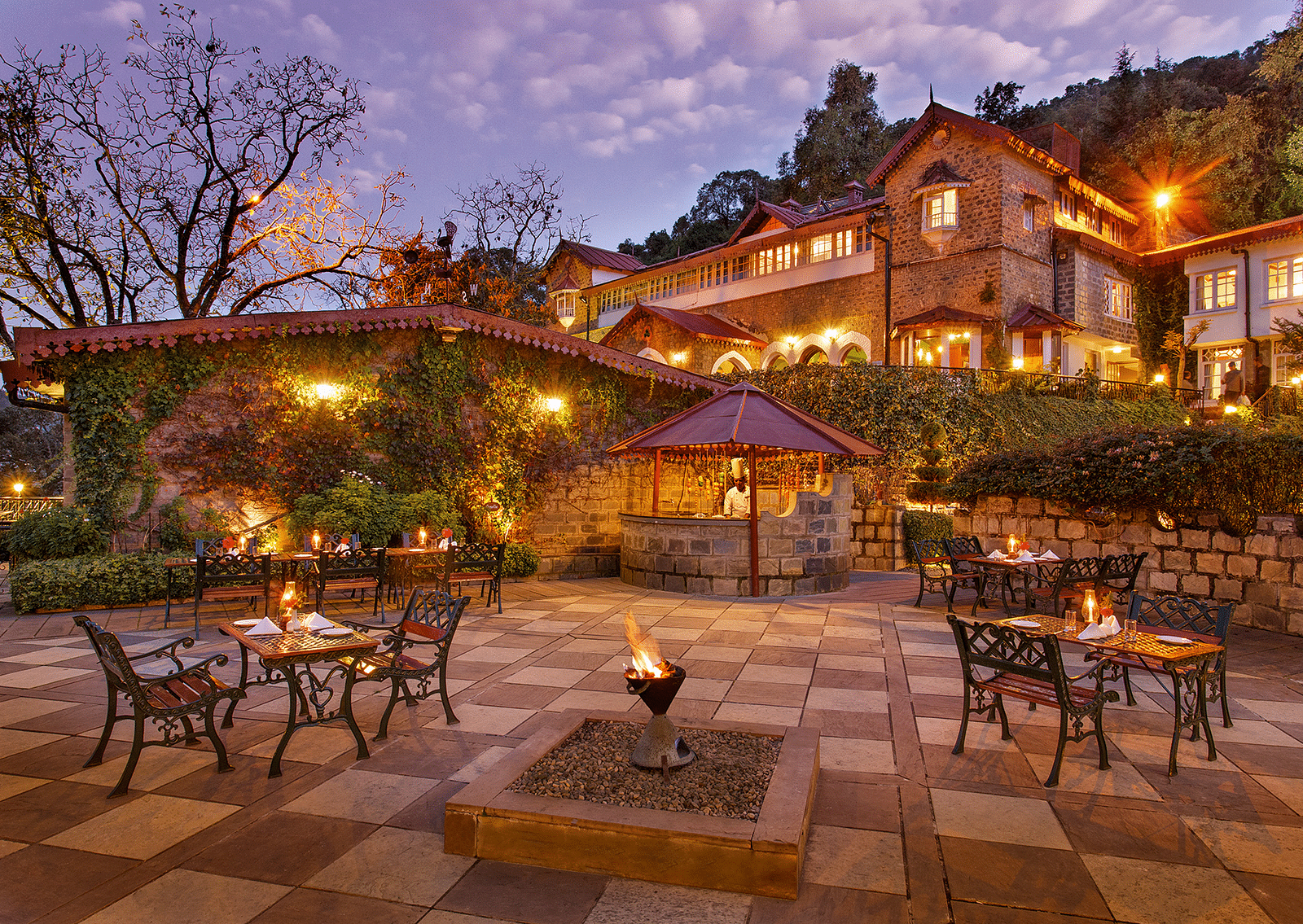A beautiful outdoor dining area at the naini retreat, nainital. There are tables and chairs set up on a stone patio, with a fireplace in the center. The building is lit up at dusk, creating a warm atmosphere. 