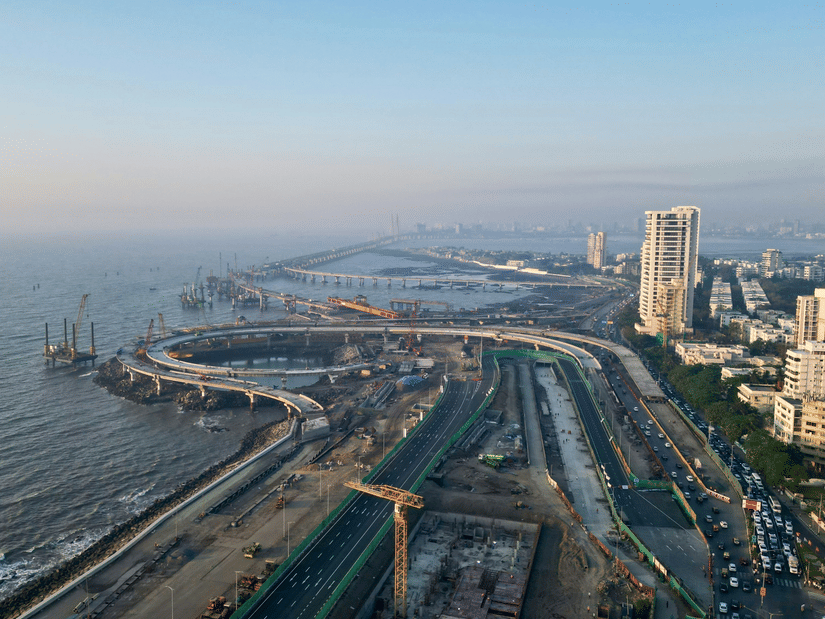 Aerial view of the Mumbai Coastal Road Project under construction, showing curved flyovers, cranes, and newly built roads along the Arabian Sea, with high-rise buildings lining the shoreline.