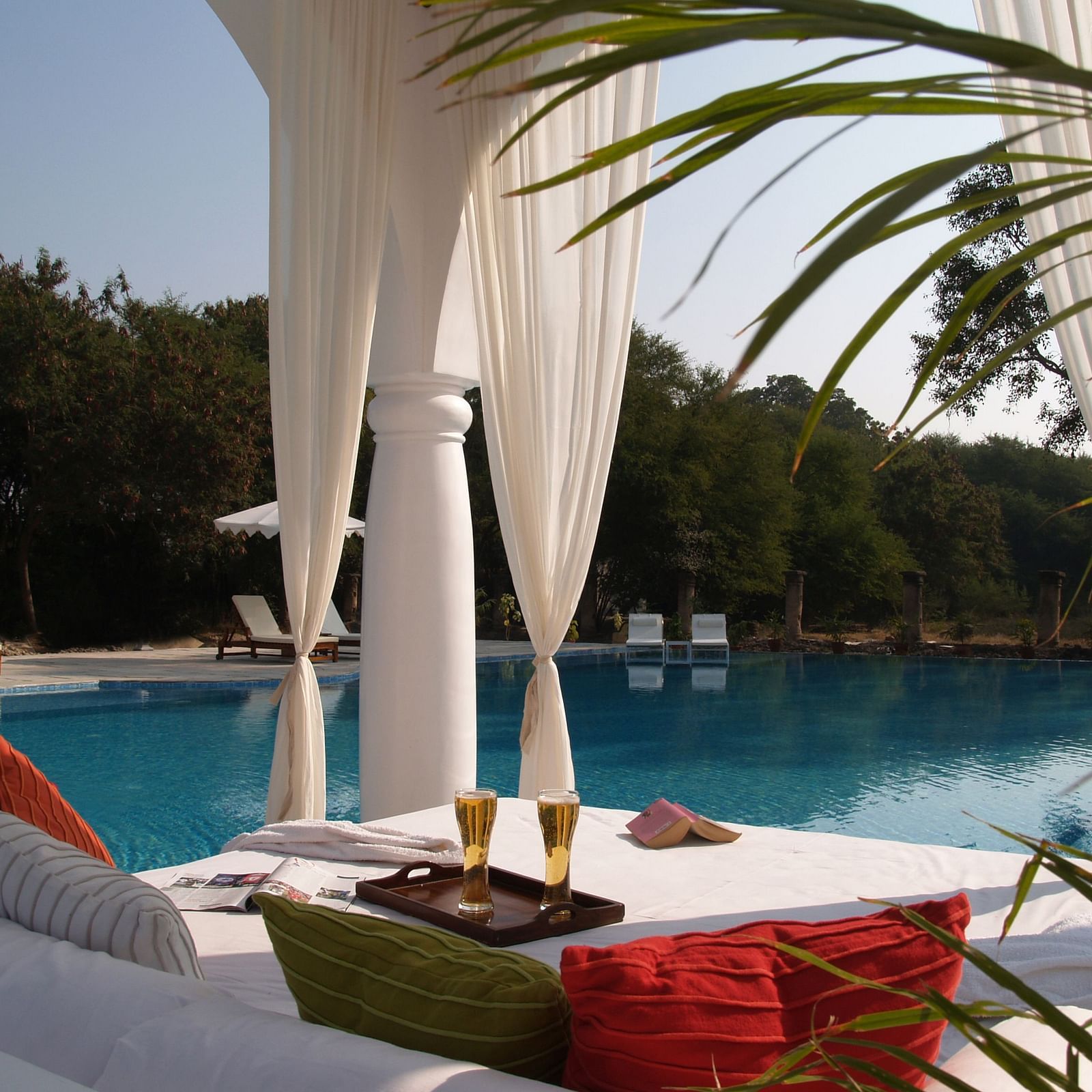 A poolside cabana with white drapes frames a view of a swimming pool, lounge chairs, and trees, with colorful pillows and drinks on a table in the foreground- Shahpura Bagh.