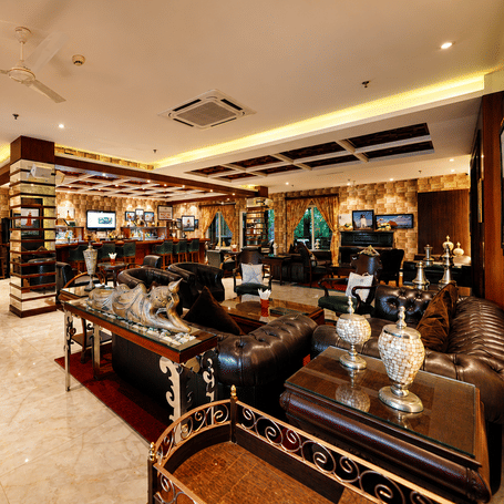 A Light House Bar MAYFAIR Palm Beach Resort, Gopalpur-on-Sea. Sofas, armchairs, and tables are arranged across the room. A bar counter with shelving stands in the background. Lighting fixtures illuminate the ceiling and walls.