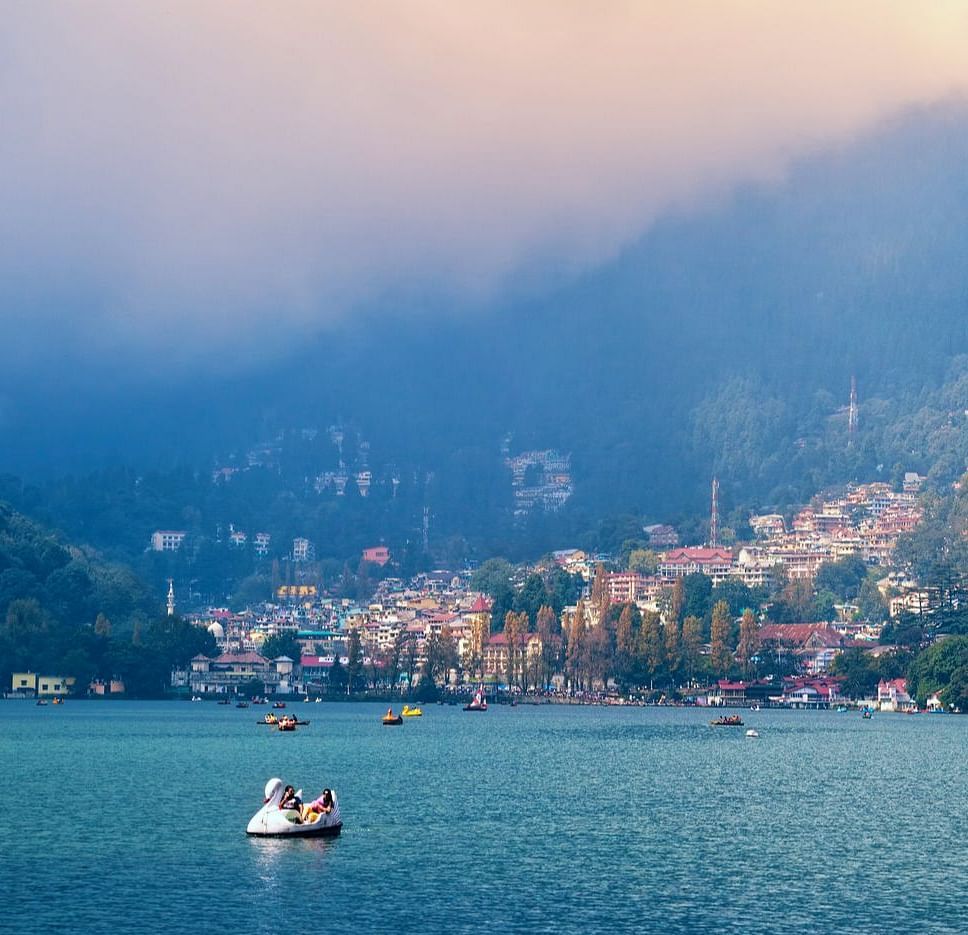 A serene view of Nainital Lake with boats floating on calm waters, backed by colourful hillside houses under a soft, cloudy sky.