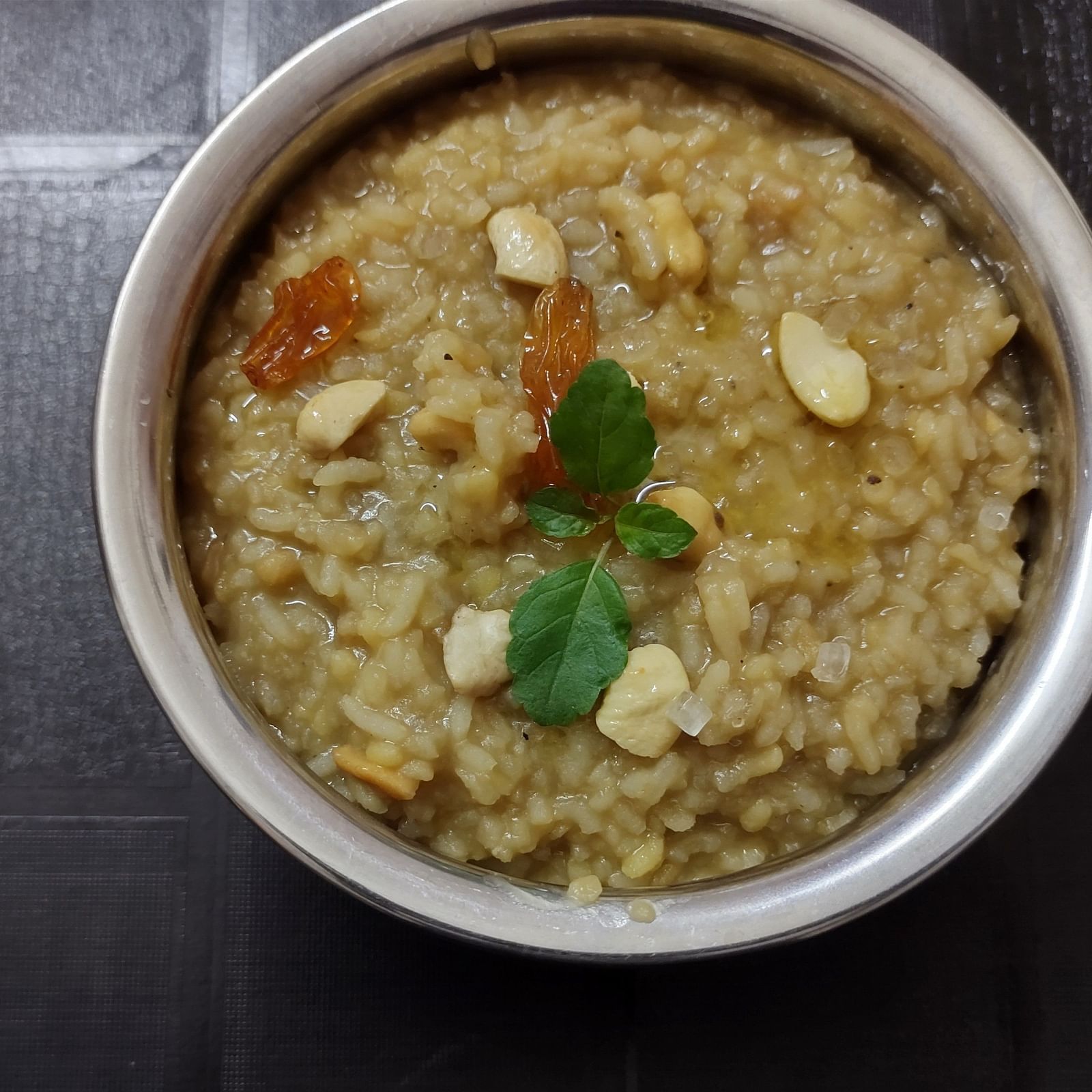 A metal bowl of a savoury Indian lentil and rice porridge, garnished with nuts, on a dark surface.