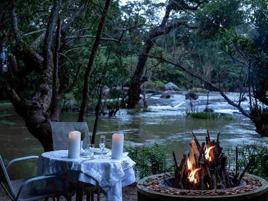 Riverside dining setup with a fire pit surrounded by lush trees at Amanvana Spa Resort, Coorg.