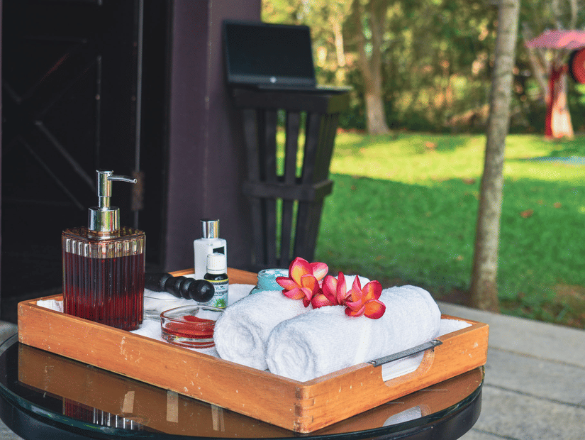 A wooden tray on a glass table holds two rolled white towels topped with pink flowers, alongside bottles and a soap dispenser, with a grassy lawn in the background.