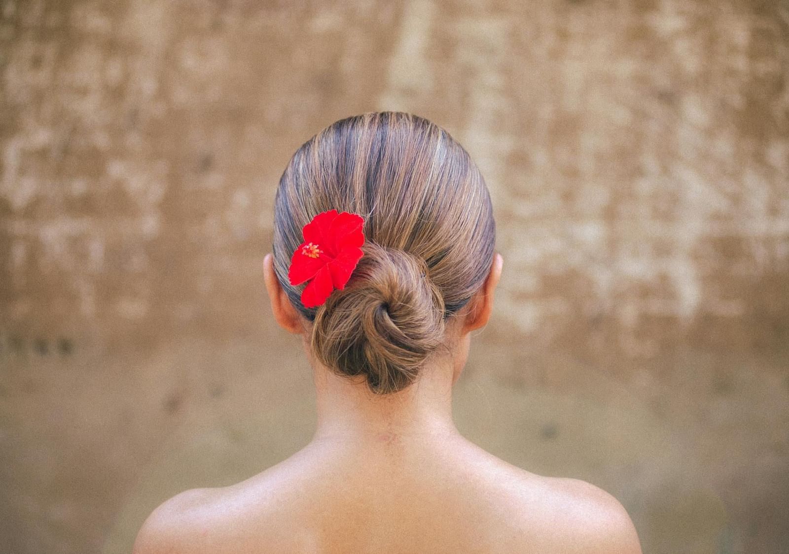 A lady with her back to the camera, wearing an updo hairstyle with a flower, facing a wall