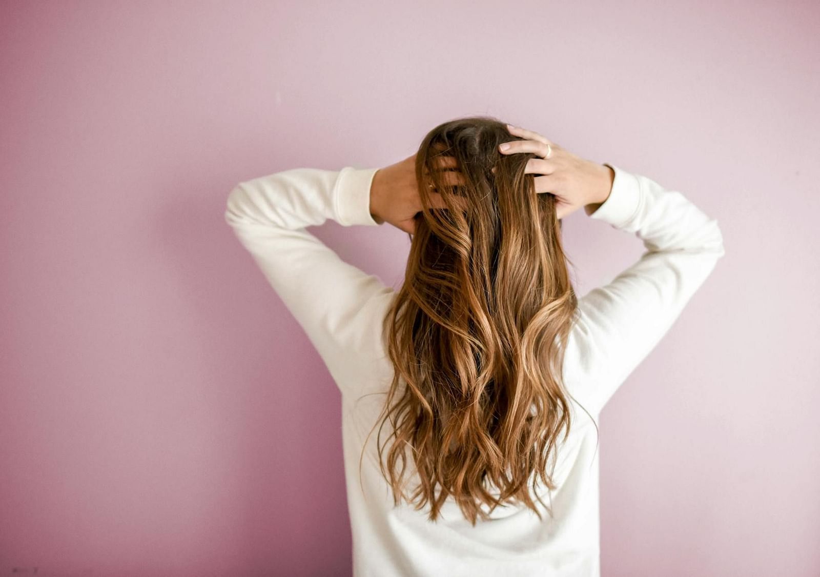 A person standing with their back to the camera, long well-styled hair, and hands behind their head touching their hair