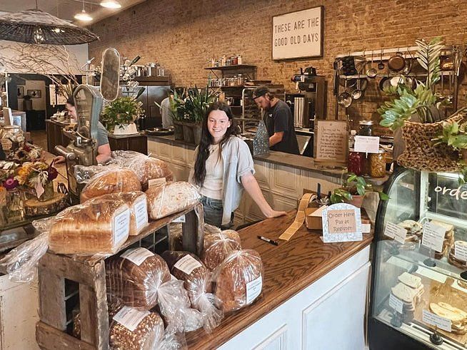 Woman standing at a counter with a big table top filled with breads ideally within a bakery and bricked wall with various objects in the background accompanied by a glass  display with light filled with breads