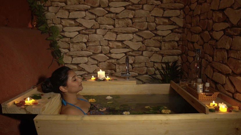A woman relaxes in a wooden bathtub filled with water and flower petals, surrounded by candles, against a stone wall backdrop in a dimly lit, spa-like setting