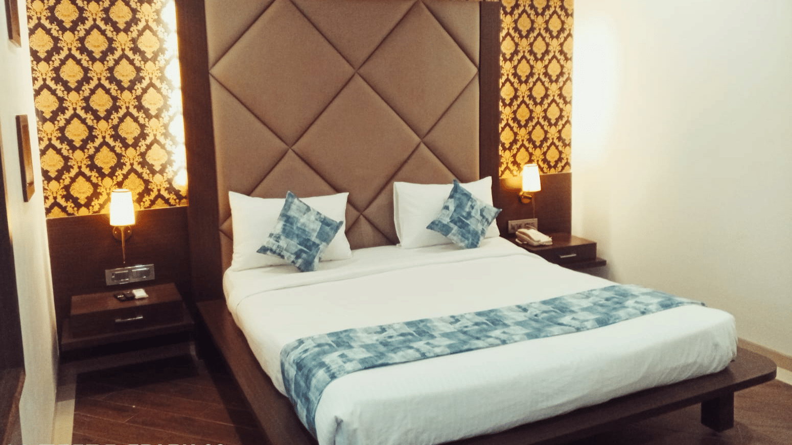 A king size bed with a padded headboard, a patterned wallpaper background, 2 bedside tables, and 2 illuminated wall lamps at Hotel Mumbai House, Thane.