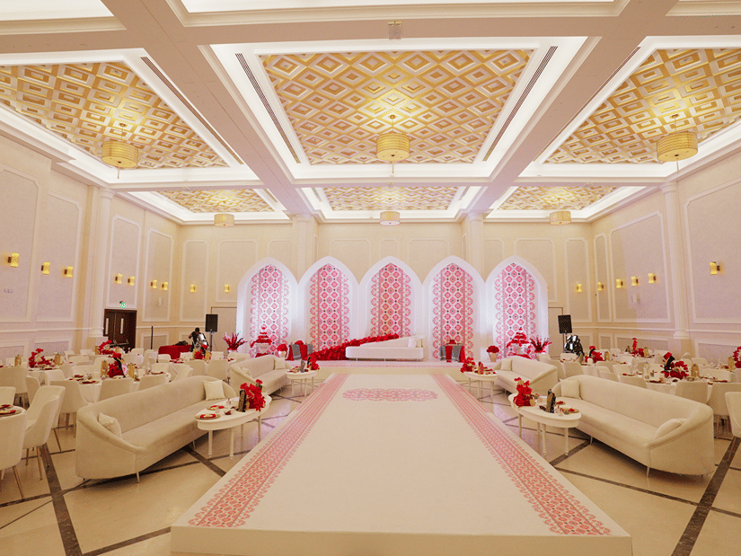 A luxurious wedding hall with elegant chandeliers, white and gold decor, and soft pink lighting highlighting an aisle lined with plush sofas. The setting is opulent and designed for an extravagant event - La Maison Hotel, Doha